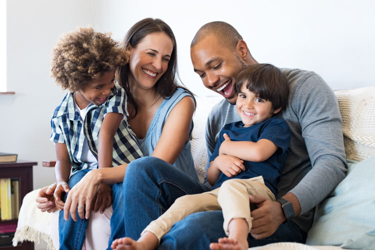 Multiracial family of mom, dad, and two sons smiles and laughs as they sit on a couch together