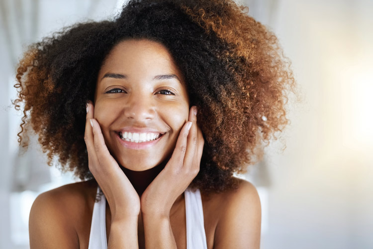 Curly-haired woman smiles with her dental implants while touching her cheeks and wearing a white tanktop