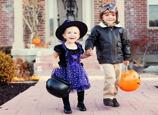 A smiling three year old girl trick or treating in a fluffy purple & black witch costume holding hands with her older brother who is wearing a bomber jacket in front of a home decorated for Halloween